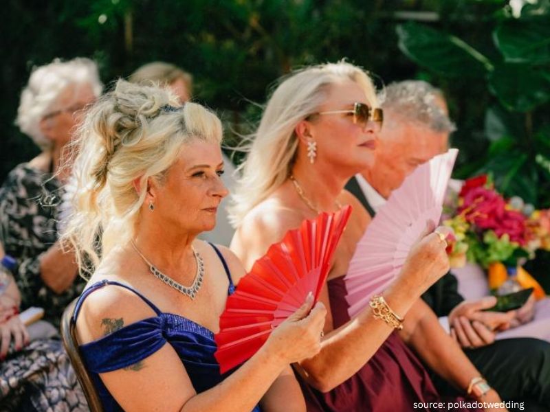 Outdoor wedding guests seated uwith fans to stay comfortable in warm weather.