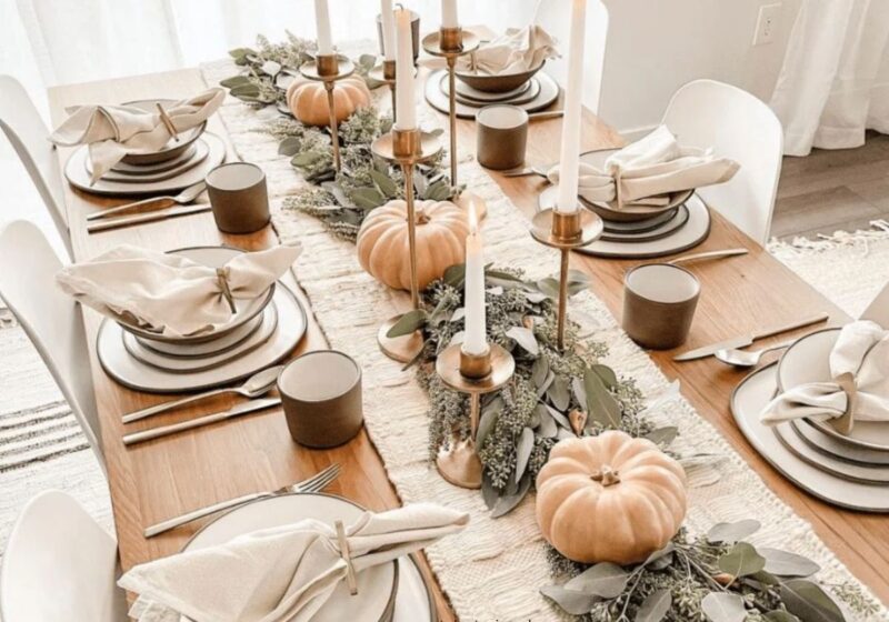 Warm Thanksgiving table with neutral linen, white plates, small pumpkins, greenery, and candles